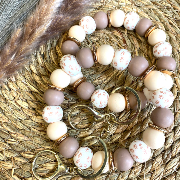 A beaded keychain bracelet with matte taupe, cream, and white speckled beads, accented with gold-toned metal rings and clasps, displayed on a woven straw mat.