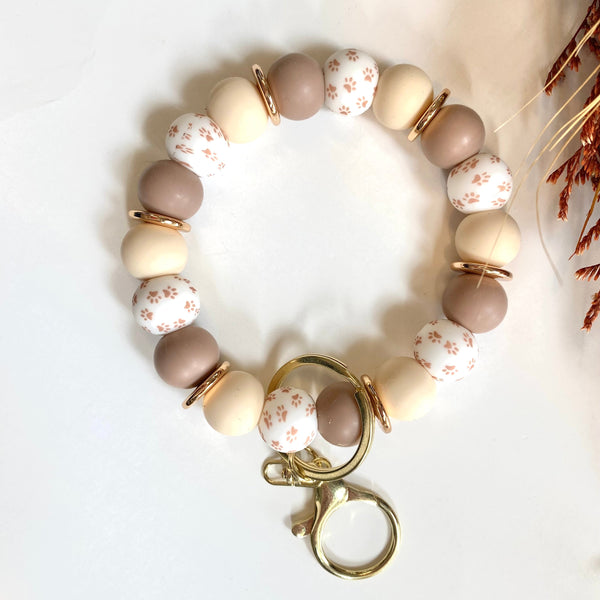 A beaded bracelet with alternating matte taupe, cream, and white floral patterned beads, accented by small gold spacer rings and a gold lobster clasp with two attached key rings, displayed on a white surface.