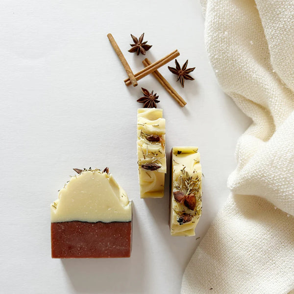 Three bars of soap with star anise and cinnamon sticks on a white surface with a beige towel.