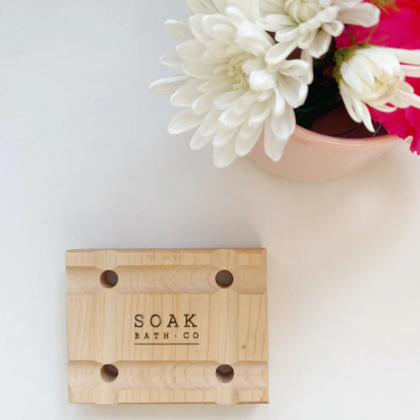 Wooden soap dish with 'SOAK BATH CO' branding on a white surface with flowers in the background.