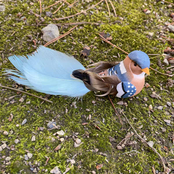 A decorative bird figurine resembling a Mandarin duck, featuring a blue cap, orange beak, patterned chest, and a large light blue tail, placed on a mossy ground outdoors.