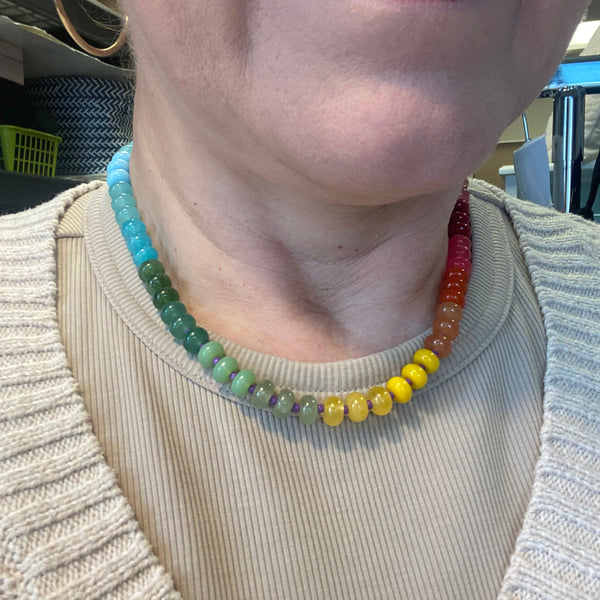 A close-up of a person wearing a beaded rainbow necklace over a beige ribbed top. The necklace features translucent round beads transitioning through light blue, green, yellow, orange, and red.
