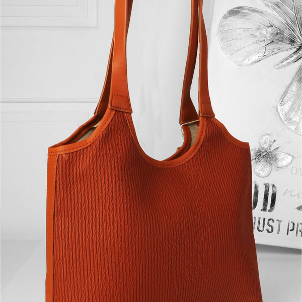An orange-brown ribbed fabric tote bag with double shoulder straps, shown on a white surface against a white wall with decorative wall art.