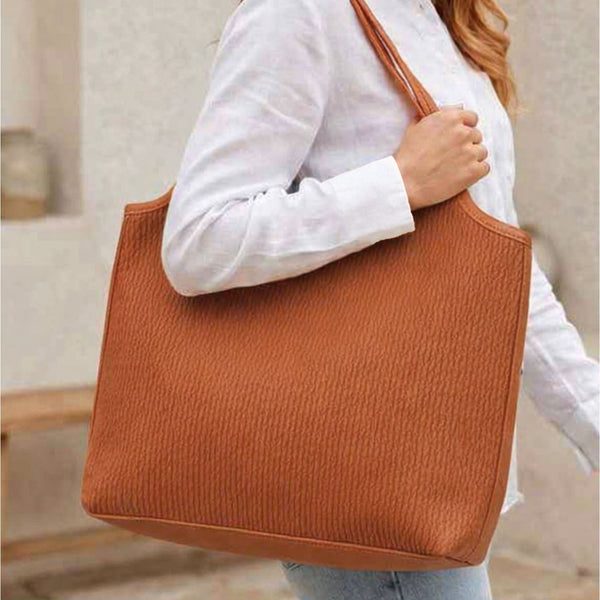 A woman wearing a white button-up shirt and light blue jeans carries a large textured brown tote bag over her shoulder in an outdoor setting.
