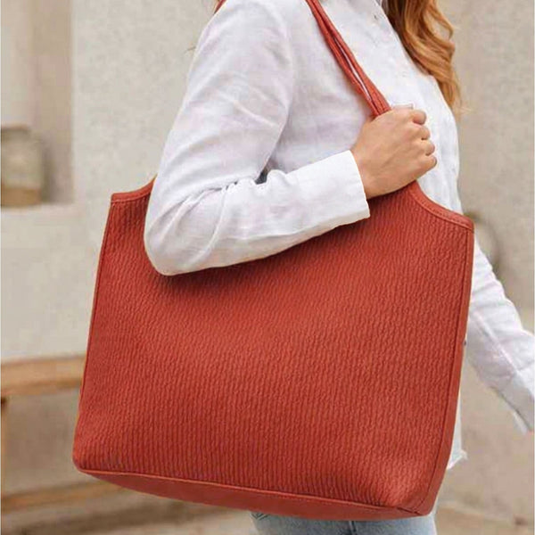 A woman wearing a white button-up shirt and light blue jeans carries a large textured terracotta orange tote bag over her shoulder in an outdoor setting.