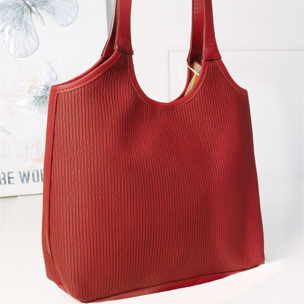 A red textured leather tote bag with long shoulder straps, displayed on a white surface against a white paneled wall with a butterfly print in the background.