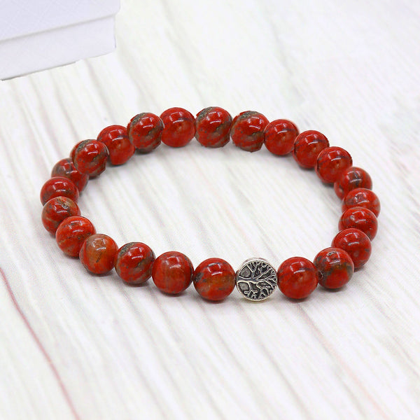 A beaded stretch bracelet made of round red jasper stones with natural brown veining, featuring a central silver-toned metal tree of life charm, displayed on a light wood surface.