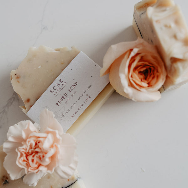 Two bars of soap with a white label and two pink flowers on a light surface
