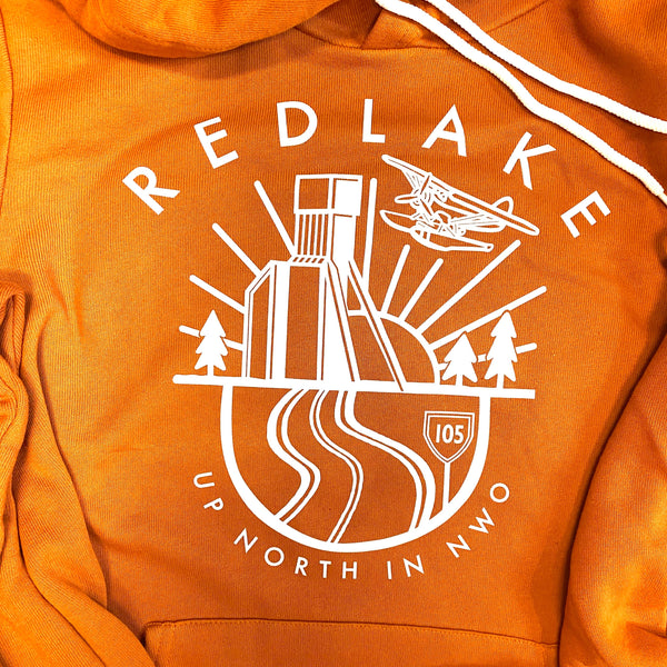 Close-up of an orange hoodie with a white graphic print on the chest. The graphic features the text 'REDLAKE UP NORTH IN NWO' surrounding an illustration of a building, trees, a road, and an airplane.