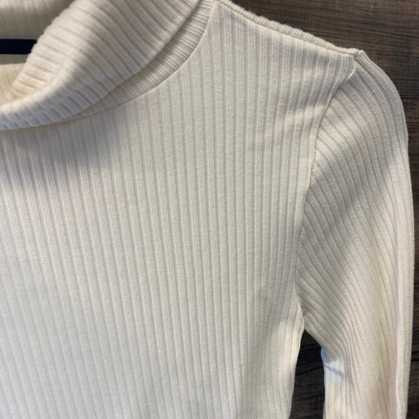 Close-up of a ribbed long-sleeve turtleneck top in off-white or cream, showing the texture and collar detail against a dark wood background.