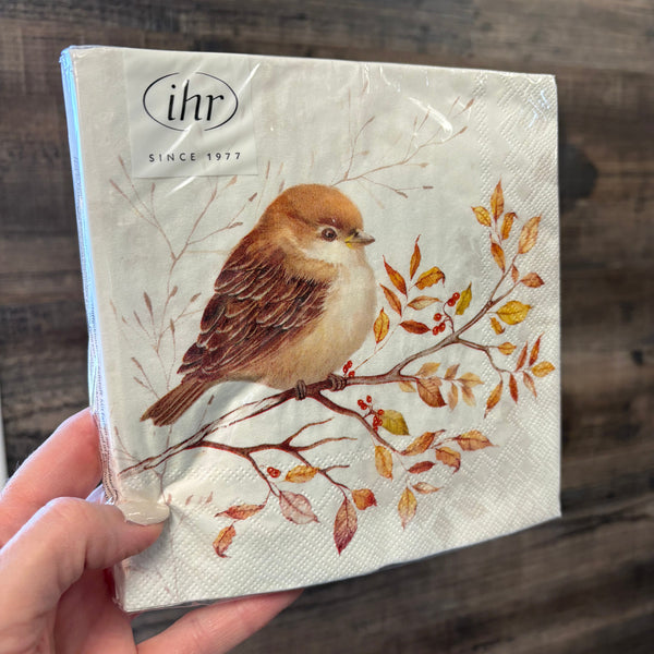 A hand holding a square paper napkin with a printed design of a small brown bird perched on a branch with autumn leaves, against a dark wood background.