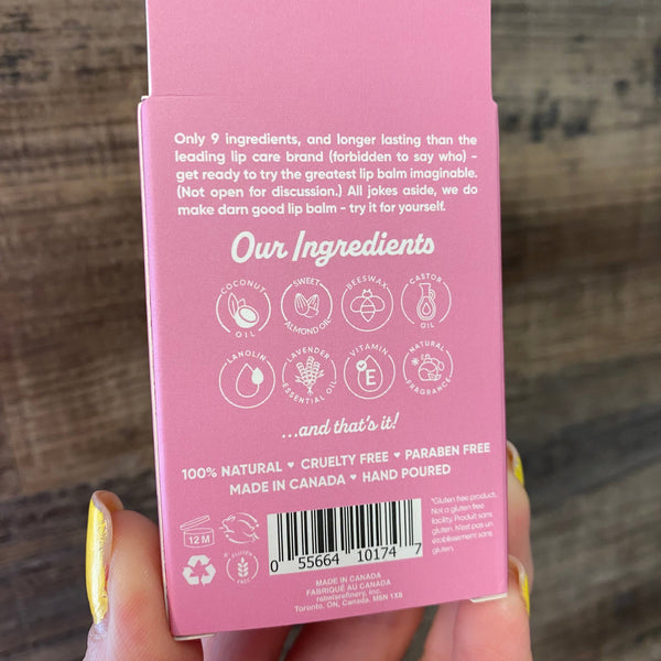 A hand with gold nail polish holds a pink cardboard box showing the back panel with product ingredients, text, a barcode, and icons, against a rustic wooden background.
