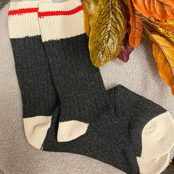 A pair of dark charcoal gray knit socks with cream toe, heel, and cuff sections, featuring a single red stripe near the cuff. The socks are laid flat on a round gray rug, styled with autumn leaves in the foreground.