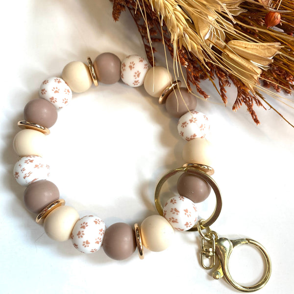 A beaded keychain bracelet with matte taupe, cream, and white floral beads, accented with gold spacer rings and a gold split ring clasp, displayed on a white background with dried wheat.