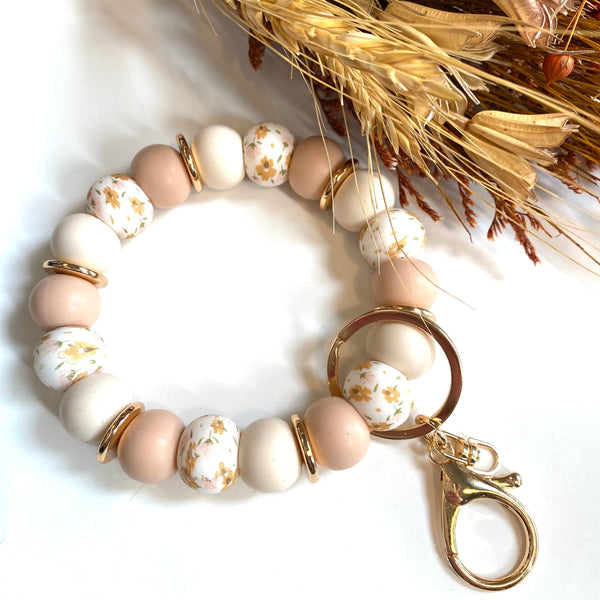A beaded bracelet with matte blush pink, cream, and white floral beads, accented by gold spacer rings and a gold lobster clasp, displayed on a white background with dried wheat stems.