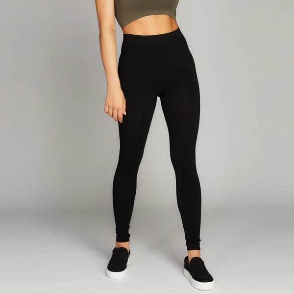 A woman modeling a two-piece activewear set consisting of an olive green sports bra and black high-waisted leggings, paired with black slip-on sneakers, against a neutral gray studio background.