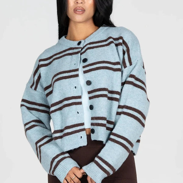 A woman modeling a cropped light blue knit cardigan with dark brown horizontal stripes and black buttons, paired with dark brown pants, against a plain light gray studio background.