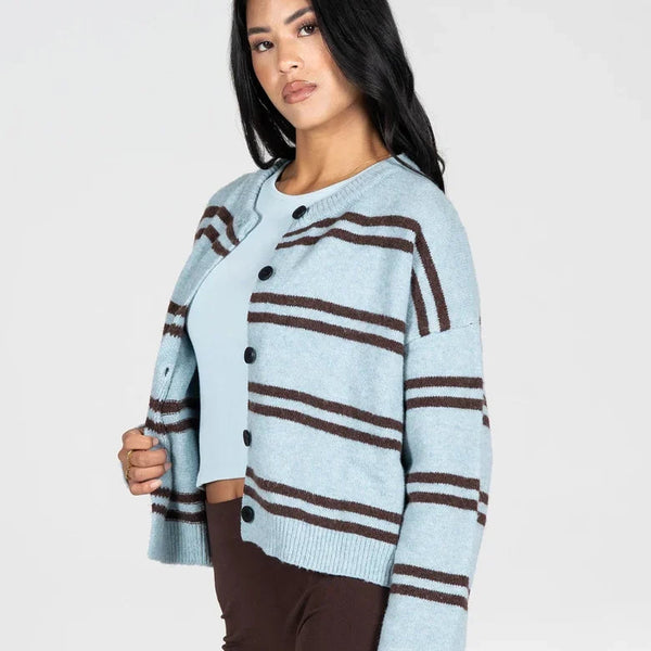 A woman modeling a light blue cropped knit cardigan with dark brown horizontal stripes, paired with dark brown pants, against a plain light gray studio background.