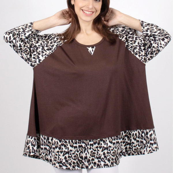 A woman modeling a brown top with leopard print sleeves and hem, paired with white pants, against a light gray studio background.