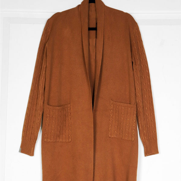A long brown open-front cardigan sweater with textured knit sleeves and two large front patch pockets, hanging against a white paneled wall.