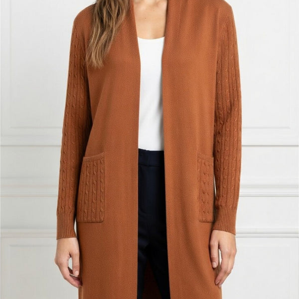 A woman wearing a rust-brown open-front cardigan with cable-knit sleeve panels and front patch pockets, styled over a white top and dark pants against a white paneled wall.