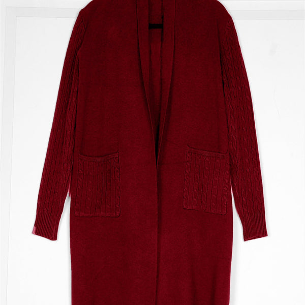 A long burgundy knit cardigan sweater with a shawl collar, front pockets, and textured cable-knit sleeve panels, hanging against a white paneled wall.