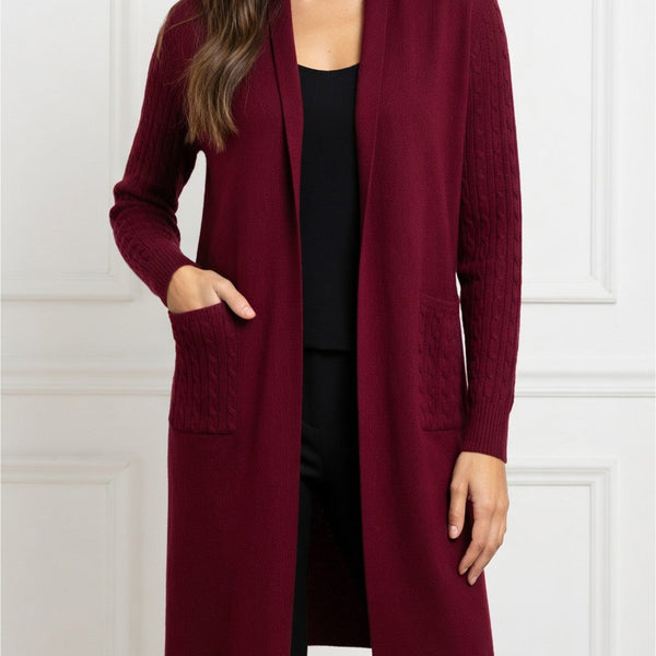 A woman wearing a long open-front cardigan in deep burgundy with textured knit sleeves and front pockets, layered over a black top and black pants, standing against a white paneled wall.