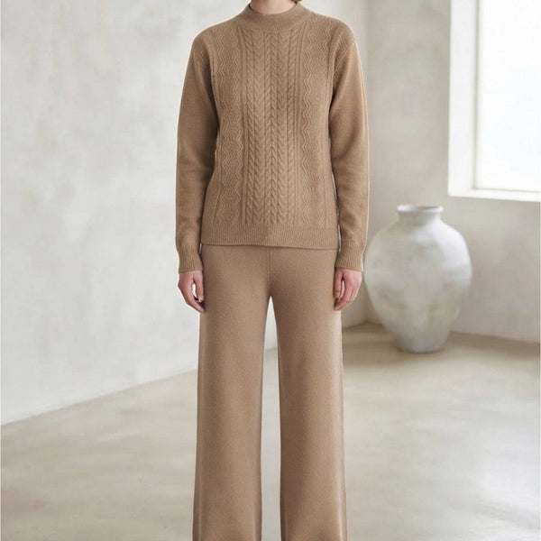 A woman stands in a minimalist room wearing a matching beige knit sweater and wide-leg knit pants set, with a large white vase near a window in the background.