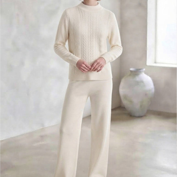 A woman stands indoors wearing a matching cream knit sweater and wide-leg pants set, with white sneakers. The room has a textured light wall, a large vase, and a bright arched window.