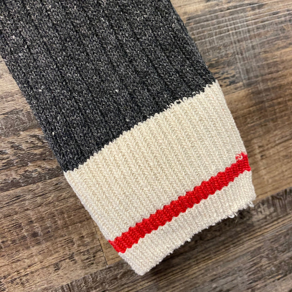 Close-up of a ribbed knit sock cuff on a wooden surface, featuring a dark charcoal gray upper section, a cream lower section, and a thin red stripe.