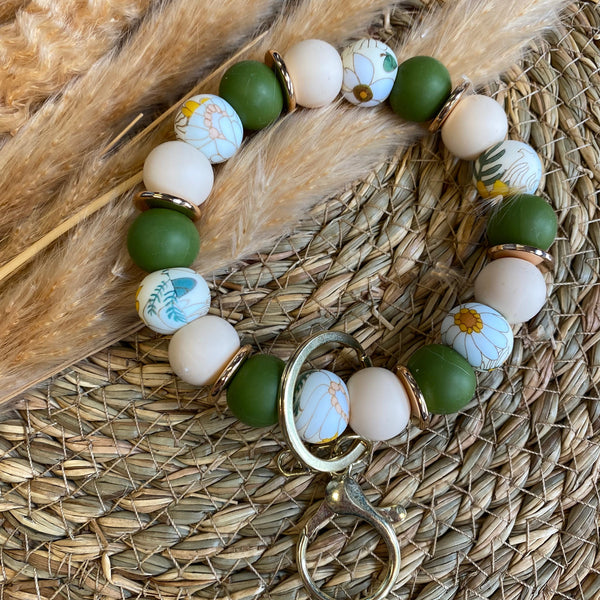 A beaded bracelet with matte green, cream, and floral patterned beads, accented by gold-toned spacers and a large gold ring clasp, displayed on a woven straw mat with dried pampas grass.