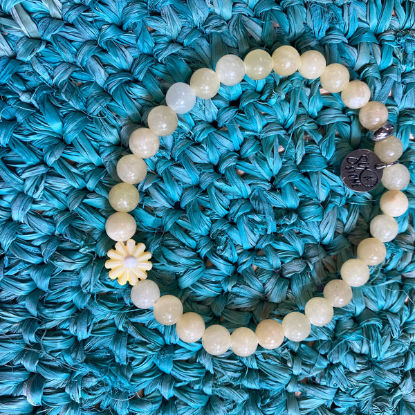 A beaded stretch bracelet made of pale cream-colored round beads, featuring a small white flower charm and a dark metallic medallion, displayed on a textured turquoise woven mat.