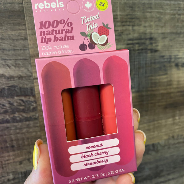A hand holding a pink box of Rebels Cosmetics 100% Natural Lip Balm in the Tinted Trio, showing three lip balms in shades of coconut, black cherry, and strawberry. The background is a dark wood surface.