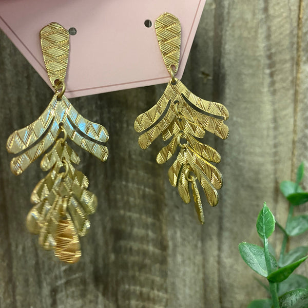 Gold leaf-shaped earrings held by a hand with red nail polish against a wooden background.