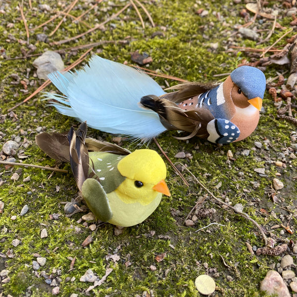 Two decorative bird figurines placed on mossy ground outdoors. One bird is bright yellow with an orange beak and olive-green wings; the other is multicolored with a blue head, brown wings, a peach chest, and a pale blue tail.