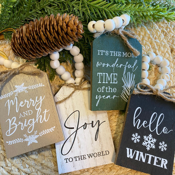 Decorative Christmas tags with festive phrases on a woven surface with greenery and pinecones.