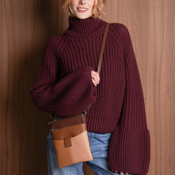 Woman wearing a maroon sweater and blue jeans, holding a brown bag against a wooden wall.