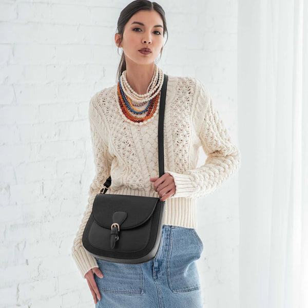 Woman wearing a cream sweater with a colorful scarf, holding a black bag against a white background