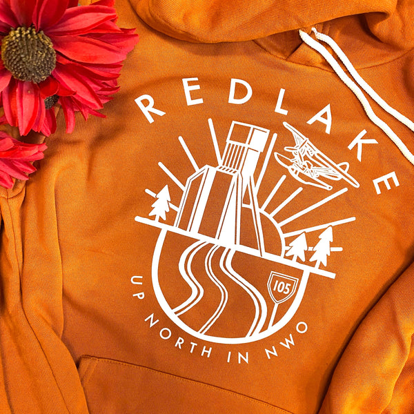 An orange hoodie laid flat with a white graphic print reading 'REDLAKE UP NORTH IN NWO' and a stylized illustration of a bridge, trees, and an airplane. A red flower is placed in the top-left corner as a prop.