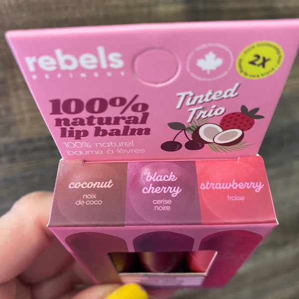 A hand with yellow nail polish holds a pink box of Rebels Refinery Tinted Trio lip balm, showing coconut, black cherry, and strawberry flavors.