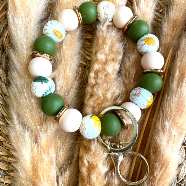 A beaded keychain bracelet featuring matte green, white, and floral-patterned wooden beads with gold-toned metal rings, displayed on a background of dried pampas grass.