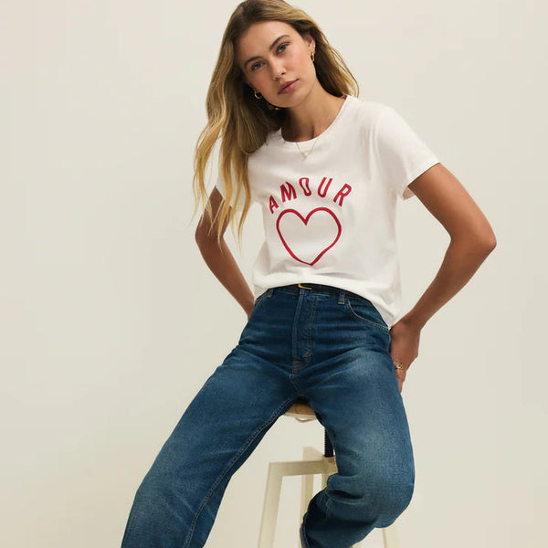 Woman wearing a white t-shirt with red text and blue jeans, sitting on a stool against a plain background.