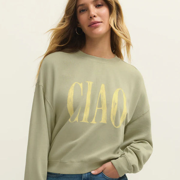 Woman wearing a green sweatshirt with 'CIAO' text and blue jeans on a beige background