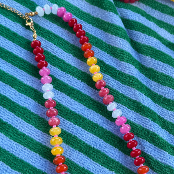 A beaded necklace with alternating strands of red, pink, yellow, and white beads, accented by a gold clasp, laid out on a green and blue striped knit fabric.