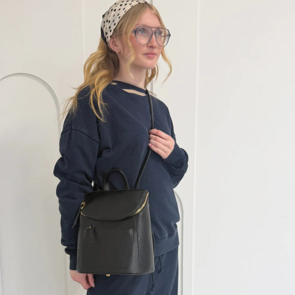 A woman wearing a navy blue sweatshirt and matching pants, a beige polka-dot headscarf, and glasses, carrying a black leather backpack with gold-tone hardware against a white wall.