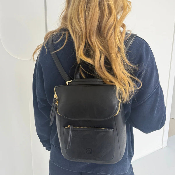 A woman viewed from behind wearing a black leather backpack with gold-tone hardware and a small circular logo on the front pocket, paired with a navy sweatshirt.