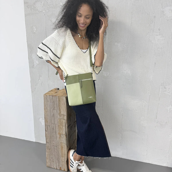 Woman holding a green and beige crossbody bag against a textured white wall.