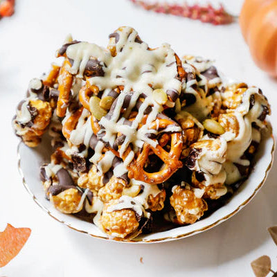 Bowl of popcorn with pretzels and chocolate drizzle on a white surface with pumpkins and leaves.