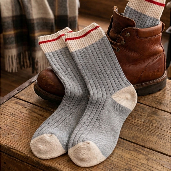 A pair of light gray ribbed crew socks with cream toe and cuff sections and thin red stripes, displayed on a rustic wooden table next to a brown leather boot.