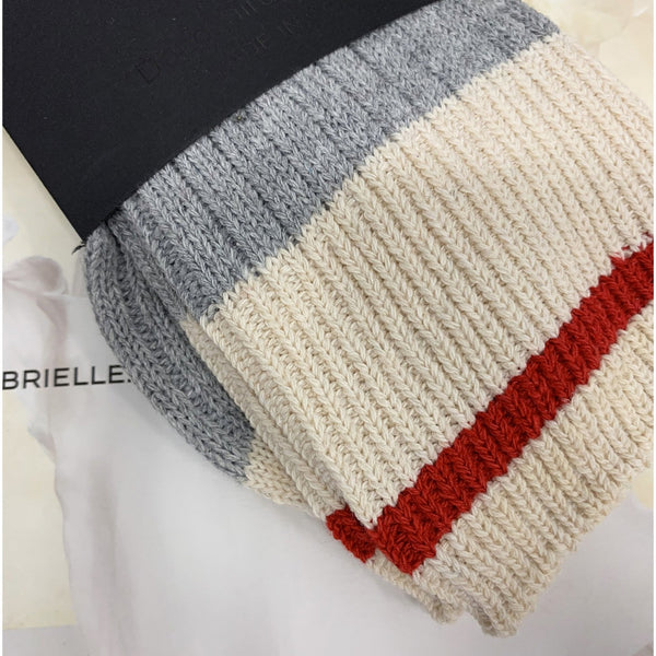A folded ribbed knit sock or bootie shown on a light surface with a black card underneath. The product has horizontal color blocks: dark gray at the top, then warm off-white/cream, with a red stripe near the lower right.